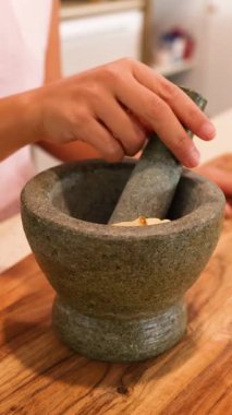 İyi aydınlatılmış bir mutfakta ahşap bir tezgahın üzerindeki baharatları öğütmek için havan ve tokmak kullanan eller.