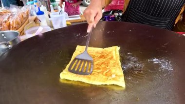Bir satıcı, Tayland pazarında parlak bir ışık altında sıcak bir ızgarada ustalıkla altın-kahverengi bir roti çevirir.
