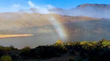 Moke Gölü üzerinde canlı bir gökkuşağı kemeri, sisli dağlar ve yemyeşil ile çevrili, parlak gün ışığında yakalandı.