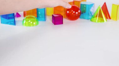 Bir el beyaz bir yüzey üzerinde renkli geometrik şekiller arasında kırmızı bir küre yuvarlar, neşeli etkileşimler ve canlı renkler gösterir