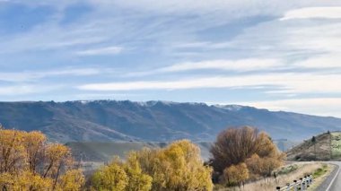 Queenstown 'un sonbahar manzarasında sakin bir yolculuk, canlı yeşillikler, uzak dağlar, ve yumuşak ışık altında berrak gökyüzü