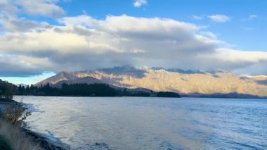 Sakin bir göl, dinamik bir gökyüzü altında heybetli dağlarla, doğal ışık altında yumuşak kamera hareketiyle yakalanmış.