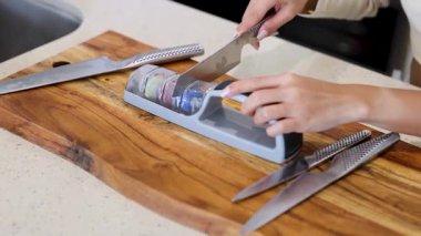 Bir insan, iyi aydınlatılmış bir mutfakta tahta kesme tahtasındaki bileme aletini kullanarak bıçağı biler.