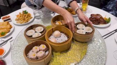 Dim sum, bambu sepetleri ve soslarla kaplı bir masa. Eller yemek için uzanıyor, canlı bir yemek atmosferi yaratıyor.