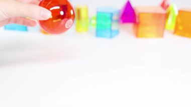 Bir el, beyaz bir yüzeyde, geometri ve renkleri vurgulayan üç boyutlu renkli şekiller arasında kırmızı bir küre ile etkileşir.