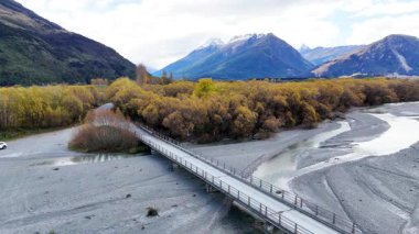Parlak gün ışığında nehir yatağının üzerinden geçen ve arkasında dağlar olan bir arabanın hava görüntüsü.