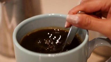 Bir el mavi bir fincanda kahve karıştırıyor, dönen desenler yaratıyor. Ilık aydınlatma sıcak mutfak atmosferini güçlendirir.