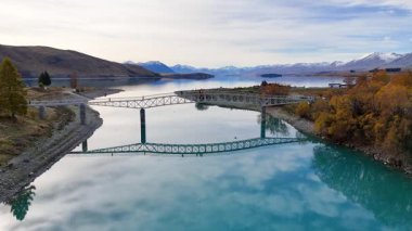 İnsansız hava aracı görüntüleri Tekapo Gölü, Yeni Zelanda üzerinde turistlerin yürüdüğü sakin bir köprü ve arka planda manzaralı manzaralar yakalıyor.