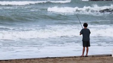 Kumlu bir plajda, bulutlu gökyüzü altında dalgalanan bir kişi, sakin bir sahil manzarası yaratıyor.