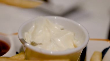 Patates kızartmasının bir fincan kremalı mayonezin içine batırıldığını gösteren bir sahne. Basit bir mutfak anını vurguluyor.