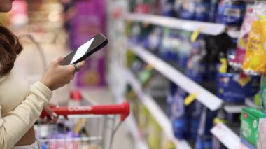Bir kadın akıllı telefonunu parlak ışıklı bir süpermarket koridorunda alışveriş yaparken kullanıyor. İçi yiyecek dolu bir arabayı itiyor.