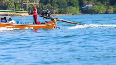 Uzun kuyruklu bir tekne huzurlu Phuket sularında yüzüyor, parlak gün ışığında canlı bir manzara ve yerel kültür sergiliyor.