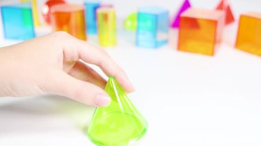 Eller, eğitici oyun ve dokunsal öğrenmeyi vurgulayarak, beyaz bir yüzeyde canlı üç boyutlu geometrik şekilleri manipüle eder.