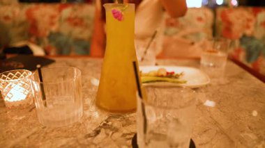 Loş bir lokanta masasına canlı bir kokteyl dökülüyor, sıcak ve davetkar bir atmosfer yaratıyor.