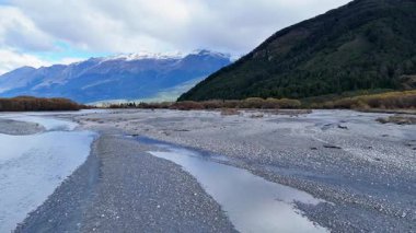Glenorchy, South Island 'da bulutlu gökyüzünün altında sakin bir nehir yatağı. Sabit kamera hareketiyle yakalanır