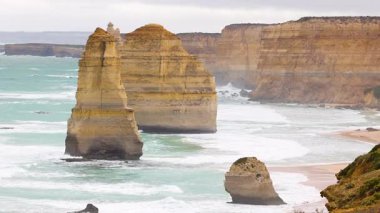 Avustralya, Port Campbell 'da bulutlu gökyüzünün altında turkuaz dalgalarla çarpıcı kıyı kaya oluşumları. Sabit kamera hareketiyle yakalanır