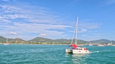 Rawai Sahili, Phuket yakınlarındaki turkuaz sularda bir katamaran süzülüyor. Açık gökyüzü ve uzak tepeler dingin bir zemin oluşturur