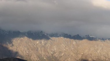 Bulutlar engebeli bir dağ sırasının üzerinden hareket ederek dinamik gölgeler oluşturuyor. Sahne değişen ışık ve atmosferik koşulları yakalar