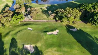 İnsansız hava aracı görüntüleri canlı yeşillikler, kum sığınakları ve parlak güneş ışığı altında sakin bir su tehlikesi içeren yemyeşil bir golf sahası yakalıyor.