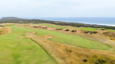 İnsansız hava aracı görüntüleri geniş golf sahasını okyanus zeminiyle, yemyeşil alanları ve yumuşak gün ışığı altında kumlu sığınakları gösteriyor.