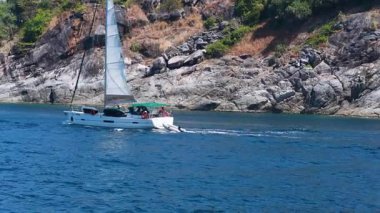 Bir yelkenli, açık mavi gökyüzünün altında Phuket 'in kayalık kıyı şeridi boyunca sakin bir okyanus seyahati sergiliyor.