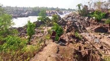 Khone Phapheng Şelalesi 'nin yanındaki kayalık yolda yürüyen bir adam, yemyeşil ve parlak gün ışığında akan suyla çevrili.