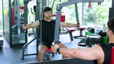 Bir adam iyi aydınlatılmış bir spor salonunda eğitmenin rehberliğinde bir makinede göğüs egzersizi yapıyor.