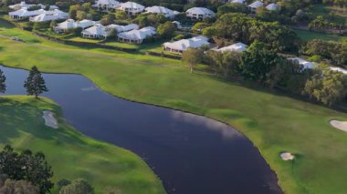 İnsansız hava aracı görüntülerinde yemyeşil, su tehlikeleri ve altın günbatımı saatinde yakınlardaki yerleşim alanlarının olduğu sakin bir golf sahası görülüyor.