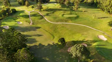 İnsansız hava aracı, verimli yeşillikler, kum sığınakları ve sıcak günbatımı ışıkları altındaki dolambaçlı yollar ile sakin bir golf sahası yakalar.