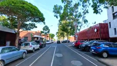 Geelong, Avustralya 'da gündüz sokak sahnesi açık gökyüzü altında park halindeki arabaları, ağaçları ve modern mimariyi gösteriyor.