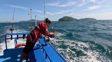 Tayland, Phuket yakınlarındaki güneşli ve berrak sularda, arka planda adalar bulunan bir tekneden bir kişi balık avlar.