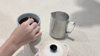 Bir el, mutfak tezgahında kaşık kullanarak şekeri kahve bardağına karıştırıyor.