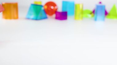 Bir el, eğitici oyun ve mekansal farkındalığı vurgulayarak, beyaz bir masa üzerinde canlı üç boyutlu geometrik şekiller düzenler.