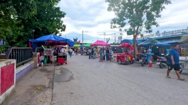 Renkli tepe örtüleri altında satıcılar ve müşterilerle canlı bir pazar sahnesi. Gündüz aydınlatma, canlı atmosfer ve çeşitli tezgahlar hareketli bir ortam yaratır.