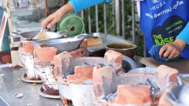 Bir satıcı kalabalık bir Phuket pazarında sıcak tavayla geleneksel Tayland atıştırmalıkları hazırlıyor. Parlak gün ışığı canlı sahneyi güçlendirir.