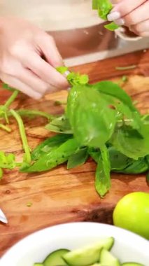 Eller ahşap bir tahtanın üzerinde taze fesleğen yapraklarını dikkatlice toplar. Parlak mutfak ışıklarıyla çevrili canlı malzemelerle.