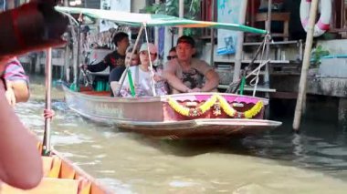 Bangkok kanal pazarında tekneler hareketli bir şekilde süzülüyor. Parlak gün ışığı satıcıları ve turistleri canlı, renkli bir sahnede aydınlatıyor.