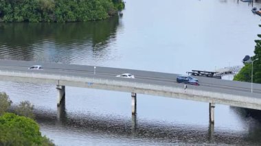 Araçlar ve yayalar sakin suların üzerindeki köprüden geçiyor, çevresi yeşilliklerle çevrili, açık gün ışığında.