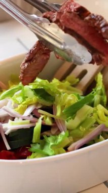 Dilimlenmiş et rengarenk bir sebze salatasına parlak bir mutfak ortamında eklenir. Tazelik ve aşçılık sanatını ön plana çıkarır.