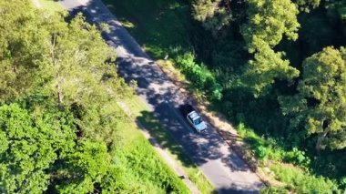 Drone, ağaçların arasından süzülen yoğun yeşillik ve güneş ışığıyla çevrili bir orman yolunda seyahat eden bir arabayı yakalar.