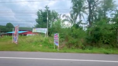 Otuz saniyelik Koh Samui yolculuğu, yemyeşil sera, yol kenarındaki binalar, ve bulutlu gökyüzünün altında bir motosikletçi