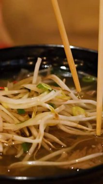 Bir insan sıcak ışıklandırmada erişte ve fasulye filizi kaldırmak için çubuk kullanan bir kase Vietnam piposu yemekten zevk alır.