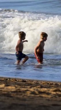 İki çocuk sahildeki okyanus dalgalarında oynamayı seviyor, parlak güneş ışığı altında, eğlence ve kahkaha anları yakalıyorlar.