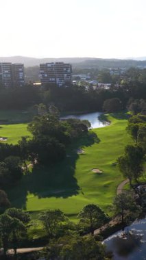 Çevresindeki ağaçlar ve uzak binalarla dolu yemyeşil bir golf sahasının gün ışığında çekilmiş hava görüntüleri.