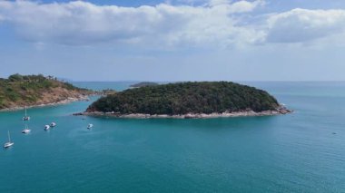 İnsansız hava aracı görüntülerinde Phuket, Tayland 'da sakin bir ada ve tekneler görülüyor. Parçalı bulutlu bir gökyüzü altında turkuaz sular ve yemyeşil alanlar sergileniyor.