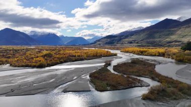 Kıvrımlı bir nehrin, dinamik bir gökyüzü altında heybetli dağlarla dolu canlı bir sonbahar yeşilliğinin havadan görünüşü.