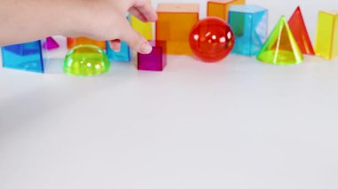 Eller, beyaz bir yüzey üzerinde renkli üç boyutlu geometrik şekillerle etkileşime girer, hareketlerini ve düzenini parlak ve oyuncu bir şekilde gösterir