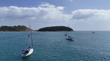 İnsansız hava aracı, sakin bir Phuket körfezinde sakin sular ve berrak gökyüzü altında yemyeşil adalar gösteren yelkenlileri yakalıyor.