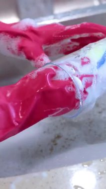 Hands in pink gloves wash a glass with soapy water in a kitchen sink, under bright lighting