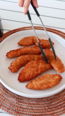 Golden-brown chicken tenders arranged on a white plate with tongs adjusting them, set on a woven placemat